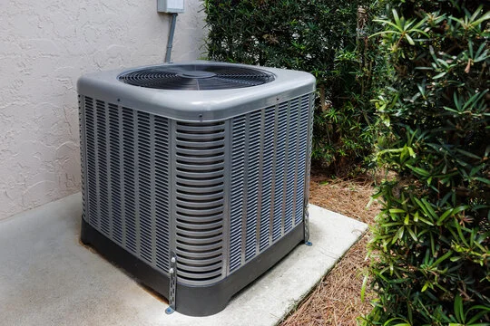 A Temperature Solutions HVAC technician inspecting an outdoor AC condenser unit at a single-family home in Lakeland, FL. The technician checks electrical connections and refrigerant lines on a sunny day with a residential neighborhood visible in the background.