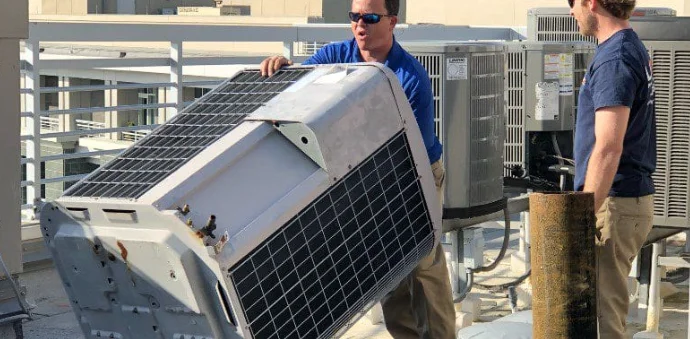 A licensed HVAC technician from Temperature Solutions installing a new outdoor AC condenser unit at a single-family home in Lakeland, FL. The replacement unit sits on a concrete pad beside the home with ductwork connections visible at the exterior wall.