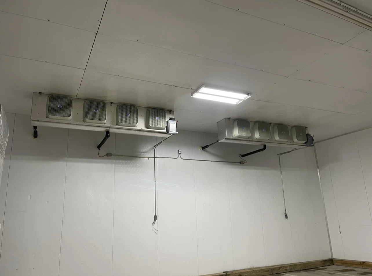 A commercial walk-in cooler unit installed inside a Lakeland, FL food service facility, with visible condenser lines and a closed insulated door. A refrigeration technician in a work uniform checks the control panel during the final commissioning step.